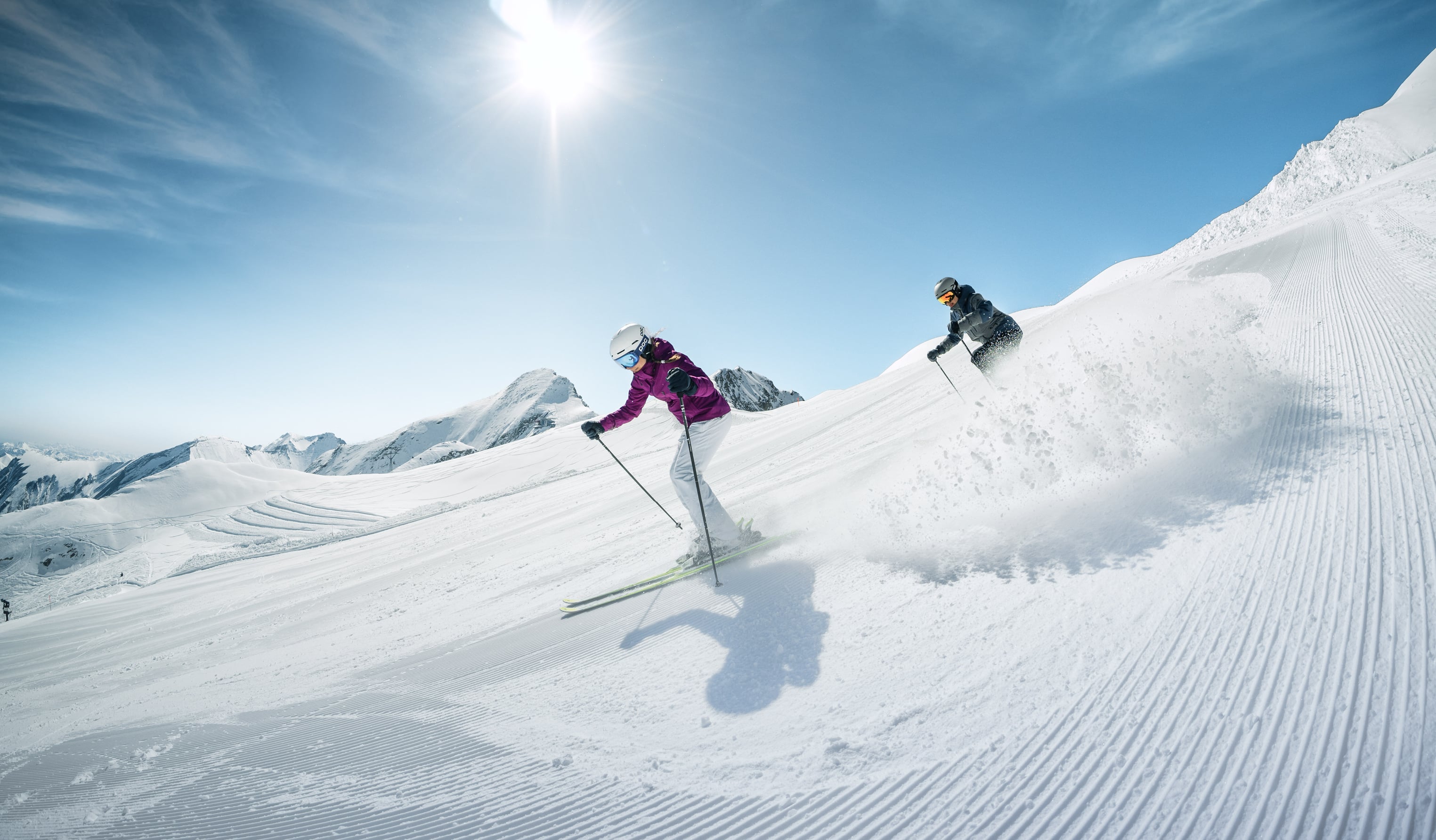 Two skiers on a groomed piste under a bright sun